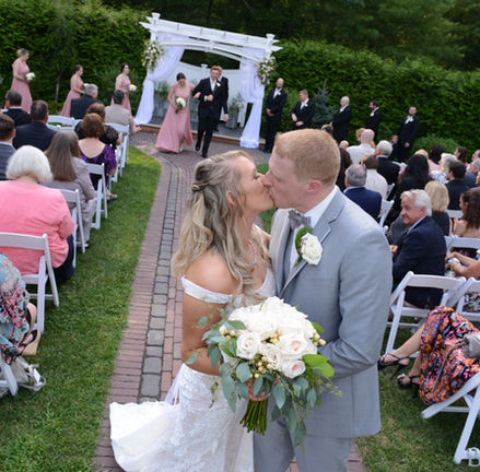 Bride and groom exchange a kiss at the end of the aisle showing the outdoor wedding ceremony location at Miraval Gardens in East Bridgewater MA