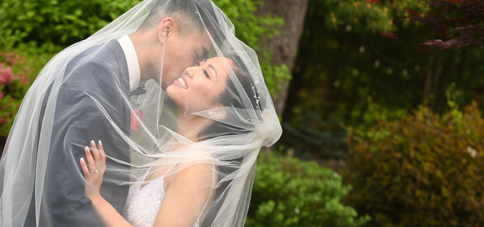 Bride is beaming with joy and love while the groom kisses her cheek under the veil with spring blooms and greenery behind them at Miraval Gardens in East Bridgewater MA