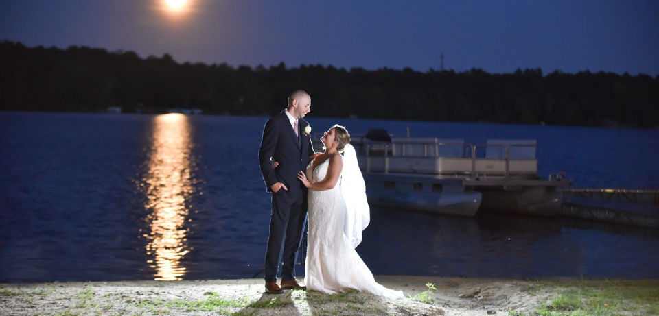 Bride and groom standing on the edge of the lake at night with the water behind them and a full moon reflecting off the water. They're backlit by flash and a second side flash on them. The camera had a long exposure to get the moon to glow.