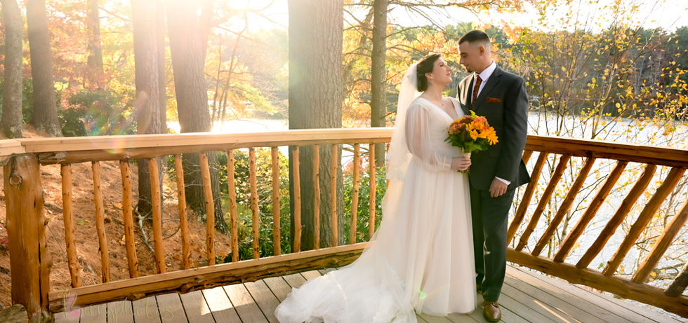 Bride and groom on the lower back deck of Camp Kiwanee in Hanson MA with the sun coming in through the trees that are in peak fall foliage and the lake behind them at Camp Kiwanee in Hanson MA