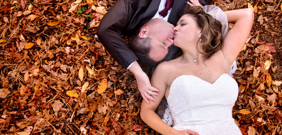 Bride and groom laying in a pile of fall leaves on their backs turning towards each other for a kiss shot from above at Loon Pond in Lakeville MA
