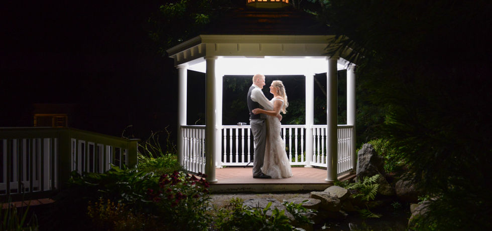 Bride and groom pose facing each other inside a gazebo at night with a single flash behind them lighting them perfectly with just enough light spilling out of the gazebo to hit the edges of the rocks, bushes and flowers just outside the gazebo while the rest of the image fades to black.