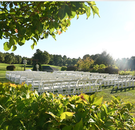 Looking through some trees and shrubs towards the outdoor wedding ceremony setup with the white chairs forming an aisle towards a floral arrangement creating a stunning scene for a wedding at Indian Pond in Kingston MA