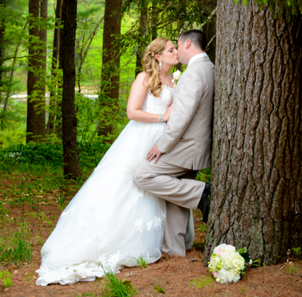 Groom leaning against a tree kissing his bride with spring and summer blooms and trees behind them at Camp Kiwanee in Hanson MA
