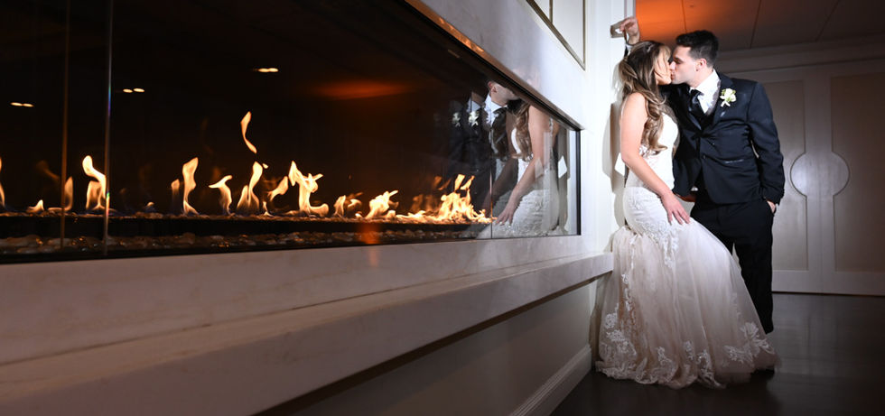 The camera is low shooting along the long fireplace with the flames dominating the entire left side of the image with the bride and groom leaning against that wall in an intimate kiss with the flash hitting only them inside the Grand Ballroom of The Villa in East Bridgewater MA