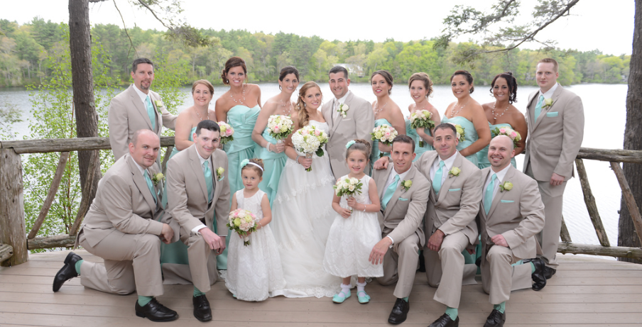 The full wedding party poses for a group shot on the back rear lower deck at Camp Kiwanee in Hanson MA with the lake and trees behind them.