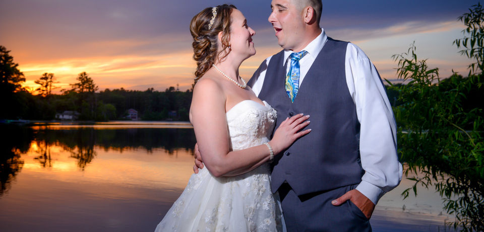 Couple poses in front of a perfect sunset with orange and purple sky reflecting off the lake at Loon Pond in Lakeville MA