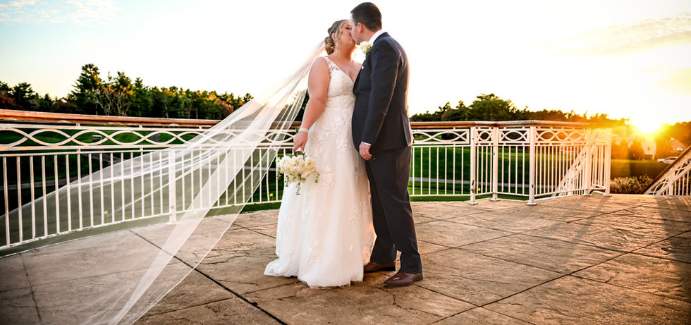 The brides vail is being swept by the wind towards the left of the image as the sun is setting to the right.  Bride and groom are locked in a kiss for a special moment alone at their wedding at Indian Pond in Kingston MA