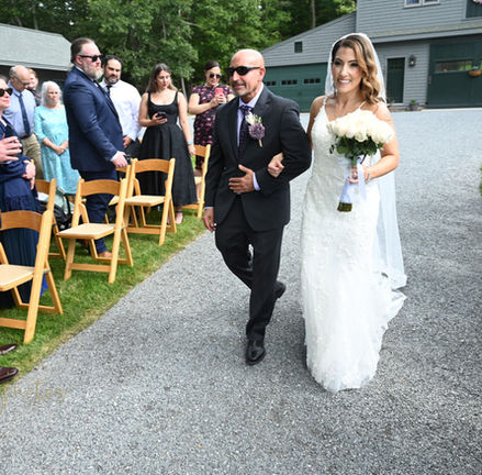 This is the the bride being escorted by her dad down the aisle at an outdoor wedding ceremony at Terrydiddle Farm in Rehoboth MA with guests on either side of the aisle and the barn as the backdrop.