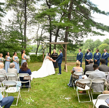 wide shot of the whole wedding with guests seated and the bridesmaids and groomsmen on either side of the bride and groom as they exchanged vows outdoors at Loon Pond Lodge in Lakeville MA