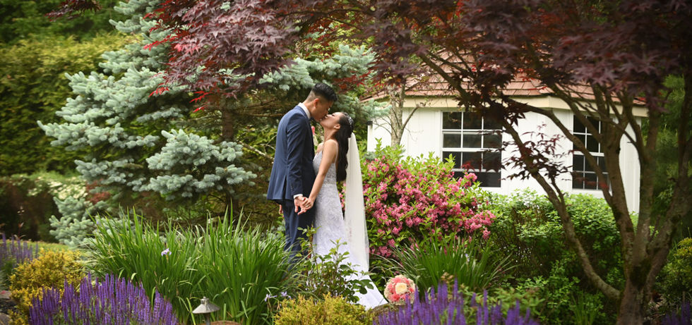 Bride and groom sneak away for a quiet moment in the gardens. This is peak color at Miraval Gardens in East Bridgewater MA with purples, greens, reds, orange, green and white florals all around them and groom in a blue suit. You've got the full rainbow naturally in the gardens at Miraval Gardens in East Bridgewater MA
