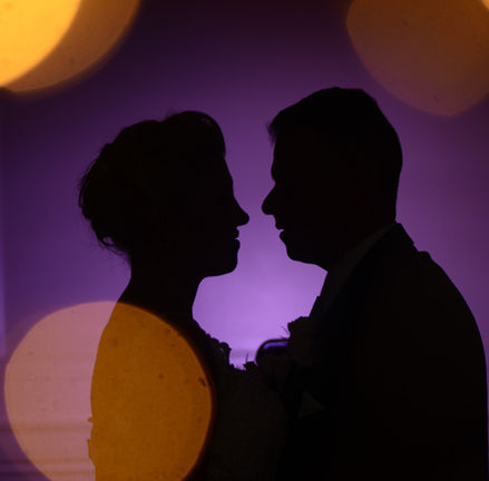Bride and groom are silhouetted against a purple uplight wall with orange bokeh balls floating around them at Granite Links in Quincy MA.  See, even if it rains you still get cool shots.
