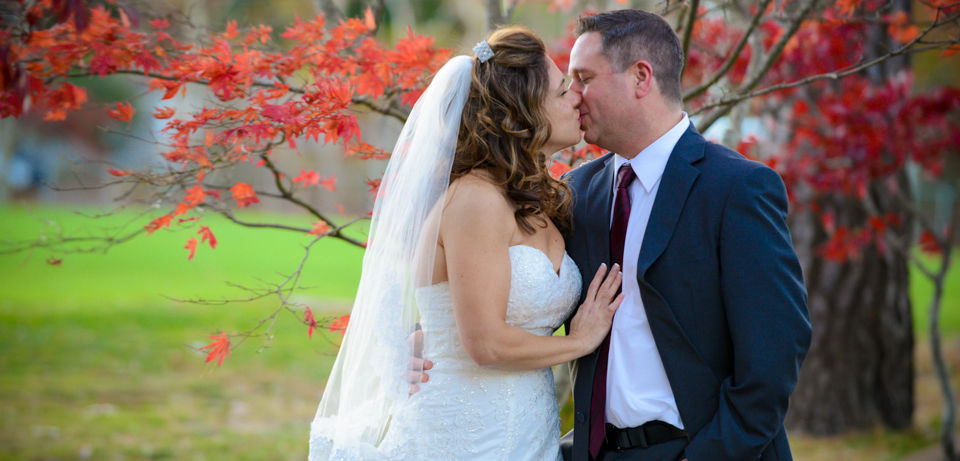 Bride and groom kiss in front of a tree in full fall foliage colors of red and orange.