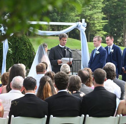 Looking through branches of a tree towards and outdoor wedding ceremony. Guests are seated in the foreground with bride and groom centered looking at each other and wedding party on either side Outdoor wedding ceremony with guests seated watching bride and groom exchange vows with bridal party on either side at Indian Pond in Kingston MA