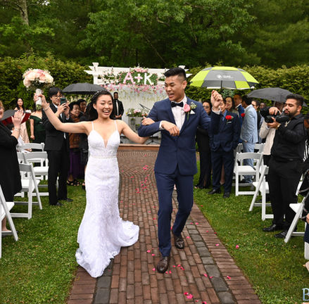Bride and groom dancing in the rain as they charge down the aisle after their ceremony with guests holding umbrellas in the rain behind them.