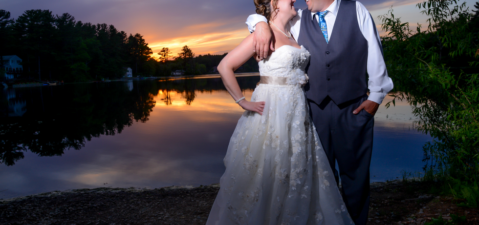 Bride and groom stand lakeside with an amazing sunset behind them and the colors reflecting off the water at Loon Pond in Lakeville MA
