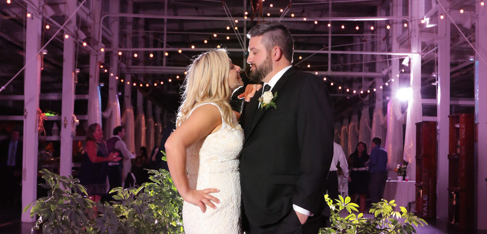 This shows the Jones River Trading Post in Kingston MA at night with purple uplighting and string lights. The bride and groom in the foreground are perfectly lit with flash.