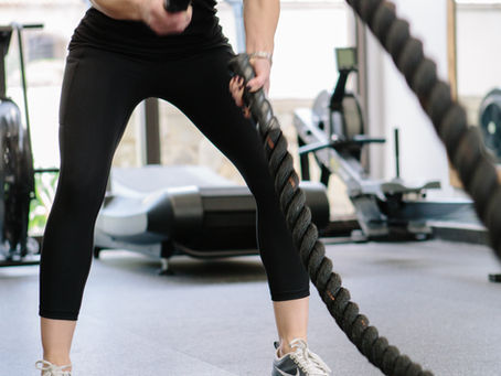 A female personal trainer at Impact One Fitness, located in Woodland Hills CA. demonstrates using battle ropes in the gym wearing black athletic wear. Bright, modern interior with exercise equipment in the background.