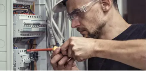 Electrician working on electrical panel, with a screwdriver, wearing safety glasses.