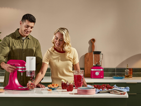 Um casal formado por uma mulher branca loira e um homem branco de cabelos escuros e curtos usando uma batedeira e um liquidificador KitchenAid