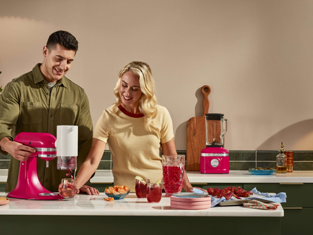 Um casal formado por uma mulher branca loira e um homem branco de cabelos escuros e curtos usando uma batedeira e um liquidificador KitchenAid