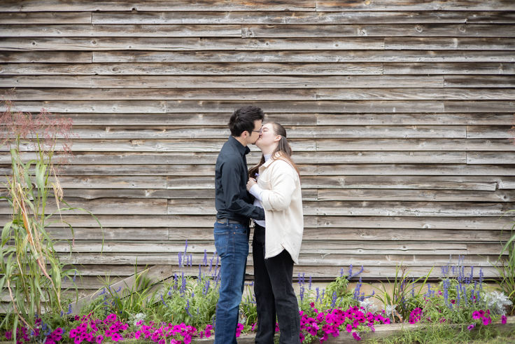Engaged couple kissing in front of a wall with weathered wood siding. There are plants, bright pink flowers, and purple flowers below. 