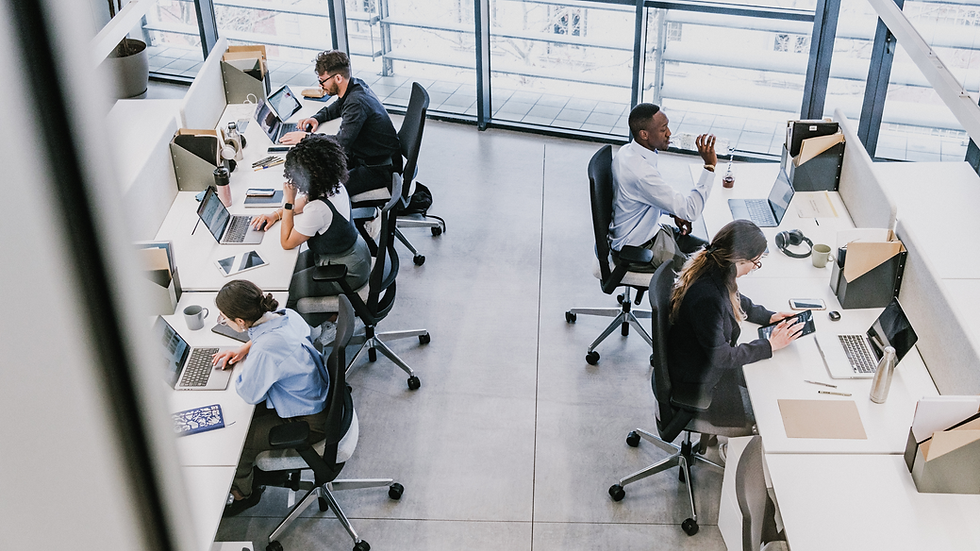 Office with people working at desks on laptops. Bright setting with large windows. Papers, tablets, and cups scattered around. Busy atmosphere.