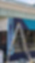 A tall ladder in front of a store window, with the outside walls of the store painted. Above the window are two diagonal strips of blue: light blue on the right, dark indigo for the left. On the left is a door, and on the right of the doorway is a vertical rectangle painted light blue on the edges. From the top to bottom is a dark indigo to light blue gradient of rectangles. At the top is a fancy light blue design built into the wall of something that looks like bells stacked on top of each other. A horizontal strip at the top of the wall is light green.