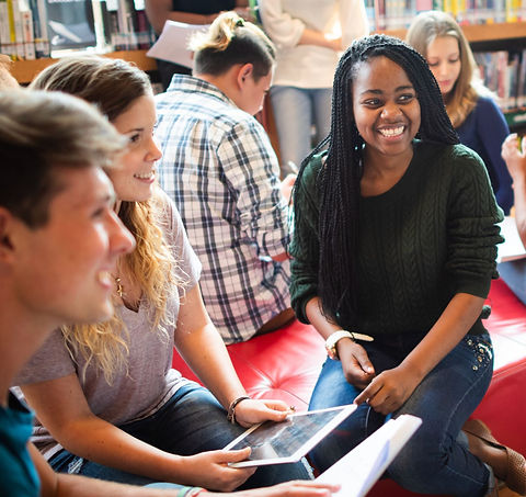 Groupe d'étudiants souriants, avec tablettes et livres, discutant dans une bibliothèque scolaire.