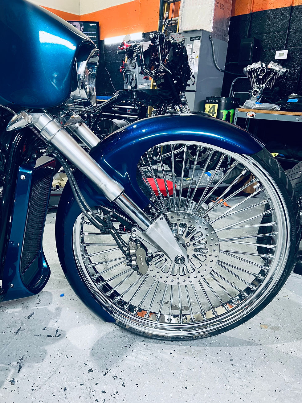 Close-up of a custom blue motorcycle front wheel with chrome spokes and brake disc in a workshop setting.