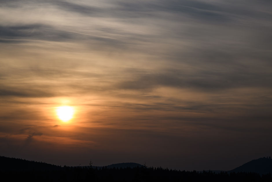 Golden sunrise through layered clouds over a dark treeline silhouette, creating a serene and moody landscape.