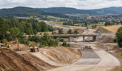Symbolbild einer Straßenbaustelle mit Erdarbeiten, Baufahrzeugen und einer Brücke in hügeliger Landschaft.