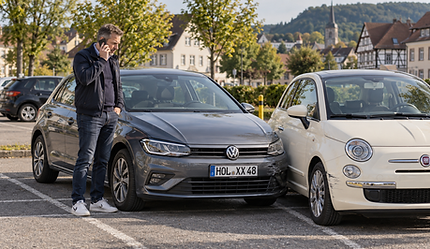 Ein Autofahrer steht mit seinem Smartphone neben zwei geparkten Autos mit einem leichten Parkschaden auf einem deutschen Parkplatz.