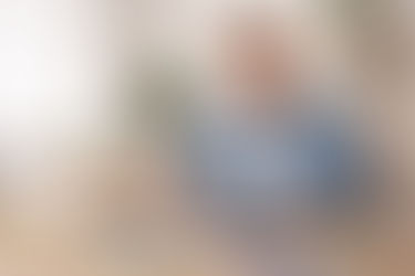 Mann in blauem Hemd lächelt am Schreibtisch mit Laptop und Modellhaus. Pflanzen im Hintergrund. Helle, freundliche Atmosphäre. Er informiert sich darüber ob er eine Energetische Sanierung steuerlich absetzen kann, trotz Förderung.