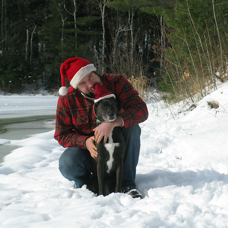 Man wearing red flannel shirt and santa hat kisses a black dog wearing a santa hat.