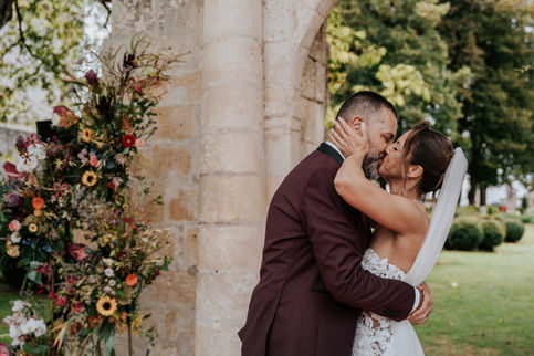 Couple de mariés s’embrassant sous une arche en pierre ornée de fleurs.
