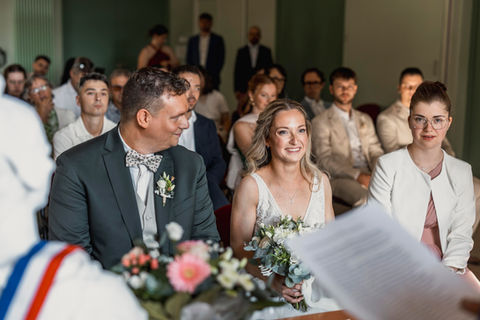 Les mariés assis à la mairie pendant la cérémonie civile, l’officiant avec écharpe tricolore au premier plan et les invités en arrière plan.