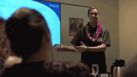 Mindfulness speaker presenting to a small audience with a slide on resilience, wearing a floral lei and gesturing to emphasize key points.