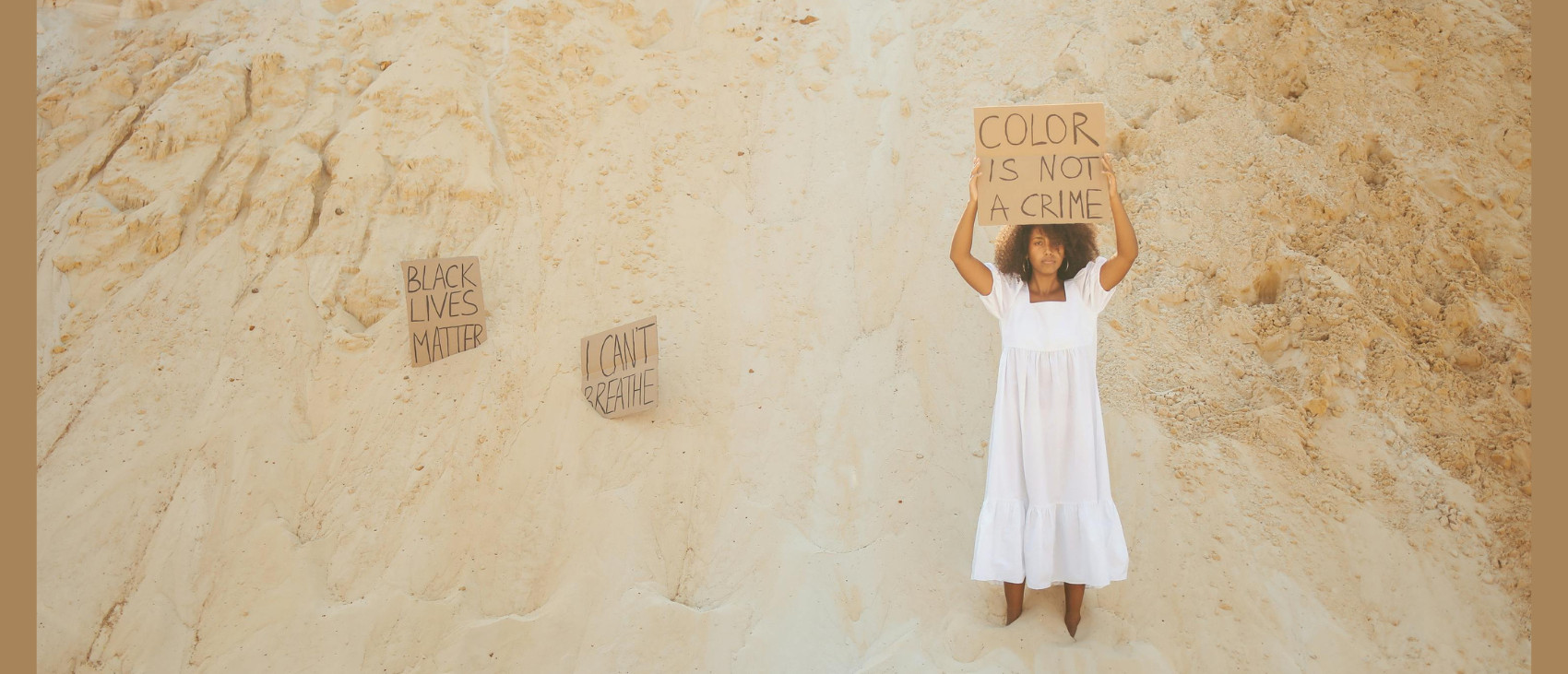 Mulher negra em um monte de areia segurando um cartaz que diz "color is not a crime". Ao lado, parcialmente enterrados na areia, outros dois cartazes: "black lives matter" e "I can't breathe!
