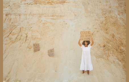 Mulher negra num banco de areia. Ela segura um cartaz que diz "color is not a crime". Na areia, estão parcialmente enterrados outros dois cartazes, que dizem: "black lives matter" e "I can't breathe".