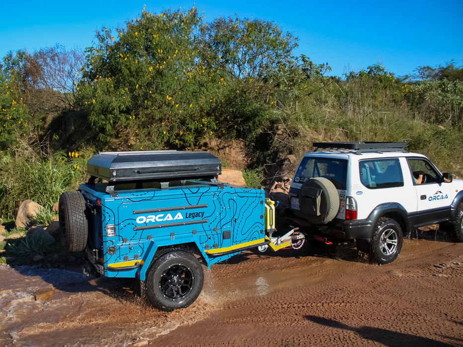Trailer being towed through a rocky river, demonstrating its rugged construction, high clearance, and off-road capability in challenging terrain.