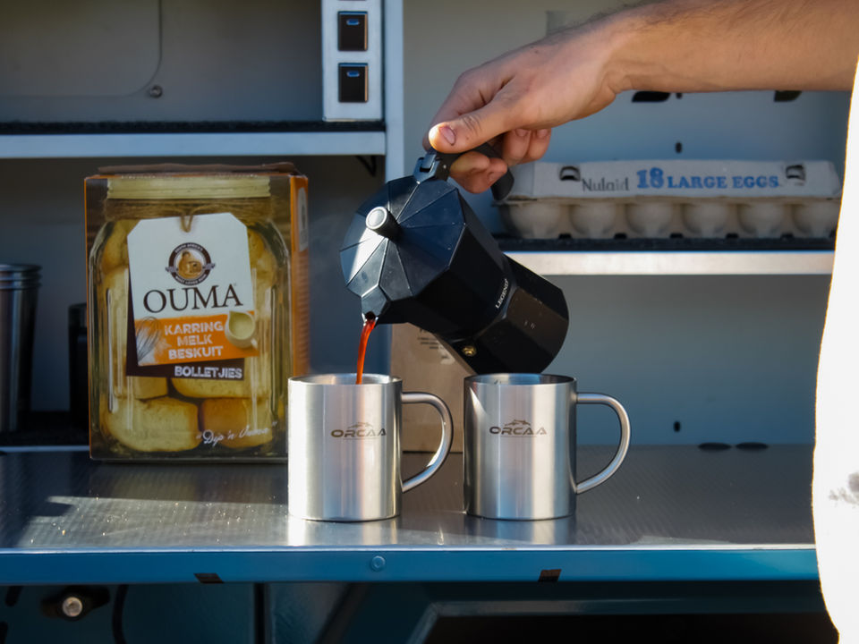 Coffee pouring from a moka pot into ORCAA-branded mugs on the trailer’s stainless steel food-safe kitchen workspace, with rusks and eggs visible in the built-in storage shelves.