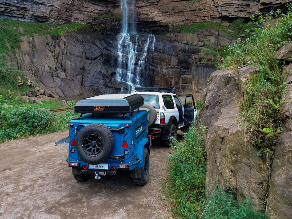 Legacy off-road trailer parked near a waterfall in a remote natural setting.