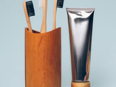 Three bamboo toothbrushes in a wooden holder alongside a metallic toothpaste tube on a light blue background.