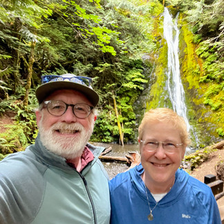 two smiling people standing in front of a waterfall, lush greenness everywhere