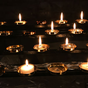 Lit candles in gold holders on a dark metal stand, glowing warmly in a dimly lit setting, creating a serene and contemplative ambiance.