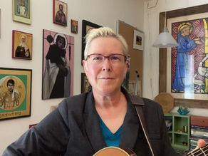 Short-haired woman with glasses holds a guitar in an art-filled room, surrounded by framed religious paintings, calm and focused