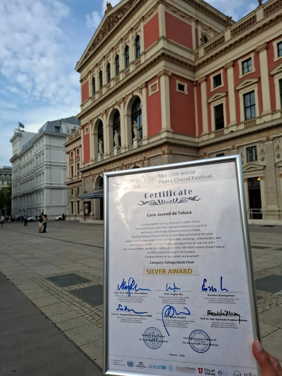 Certificado enmarcado del Coro Juvenil de Toluca, Premio de Plata, ante un edificio histórico en una plaza soleada.