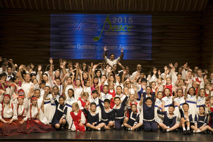 Coro infantil en escenario, niños y niñas saludan con las manos levantadas bajo cartel World Peace 2015.