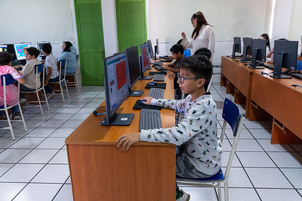 Niños en un aula de informática con una maestra; un niño usa el ordenador, mesas de madera, paredes blancas y puertas verdes.